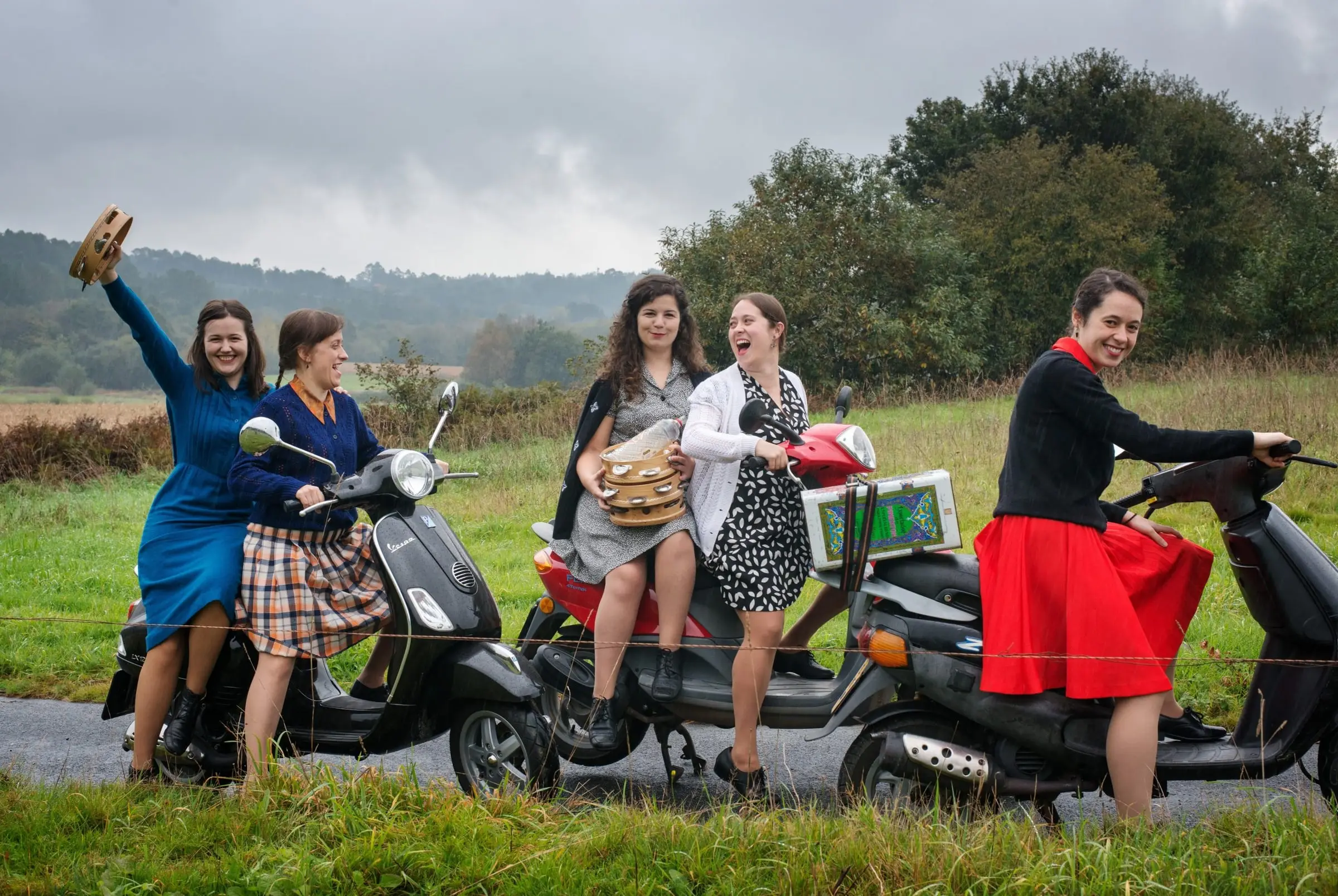 Cinco mulleres con motos nun prado; unha ergue a pandeireta